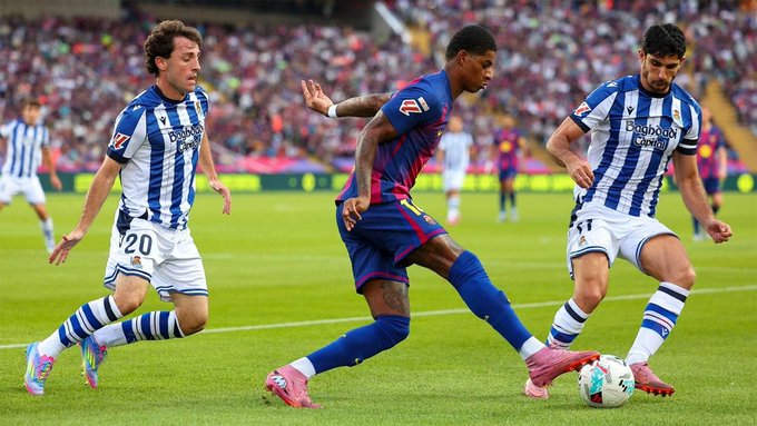 Marcus Rashford stands on a football field wearing a blue and red striped Barcelona jersey with Nike logo and club crest, black shorts, and arm tattoo visible, with stadium seating blurred in background.